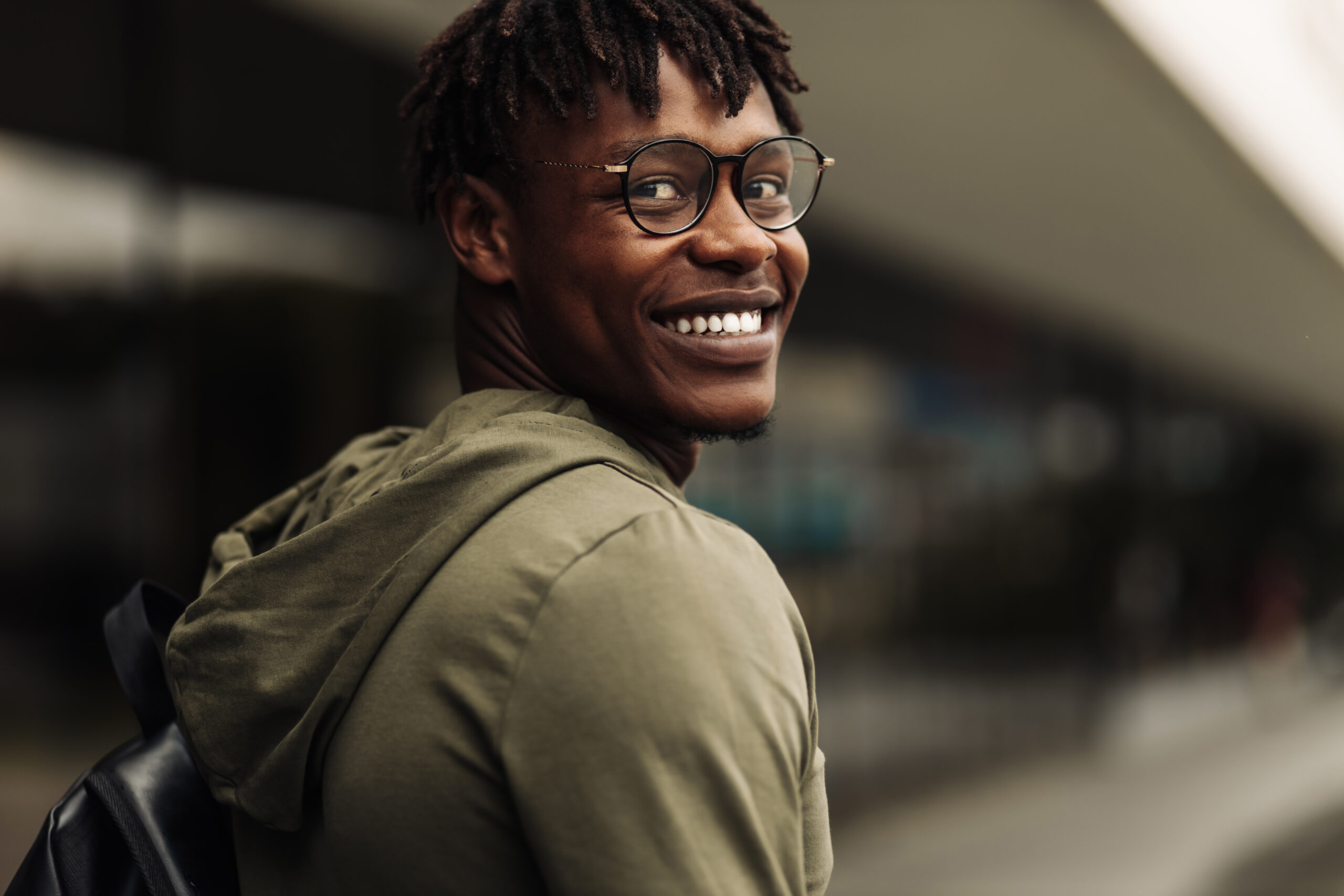 Happy handsome successful african student, wearing glasses and with a black backpack, standing on the steps and smiling, looks into the frame outdoors on the street near the university