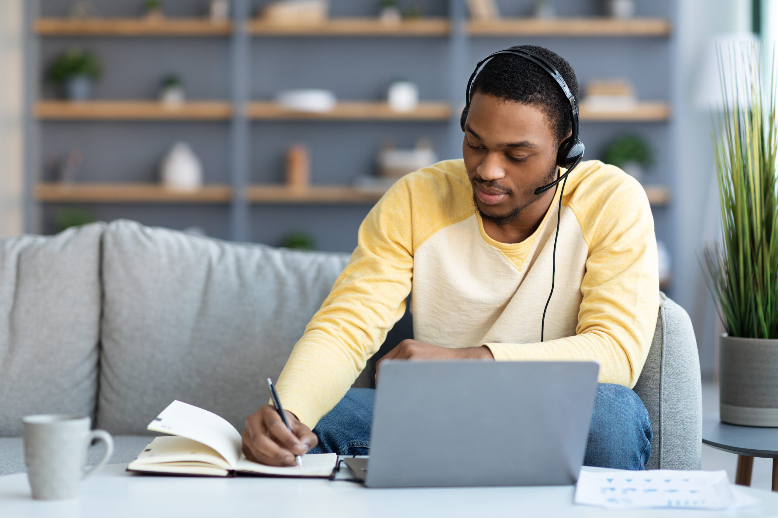 Black guy attending online training or webinar from home, sitting on couch in front of coffee table with laptop, using headset, taking notes, copy space. E-education, online class, training concept